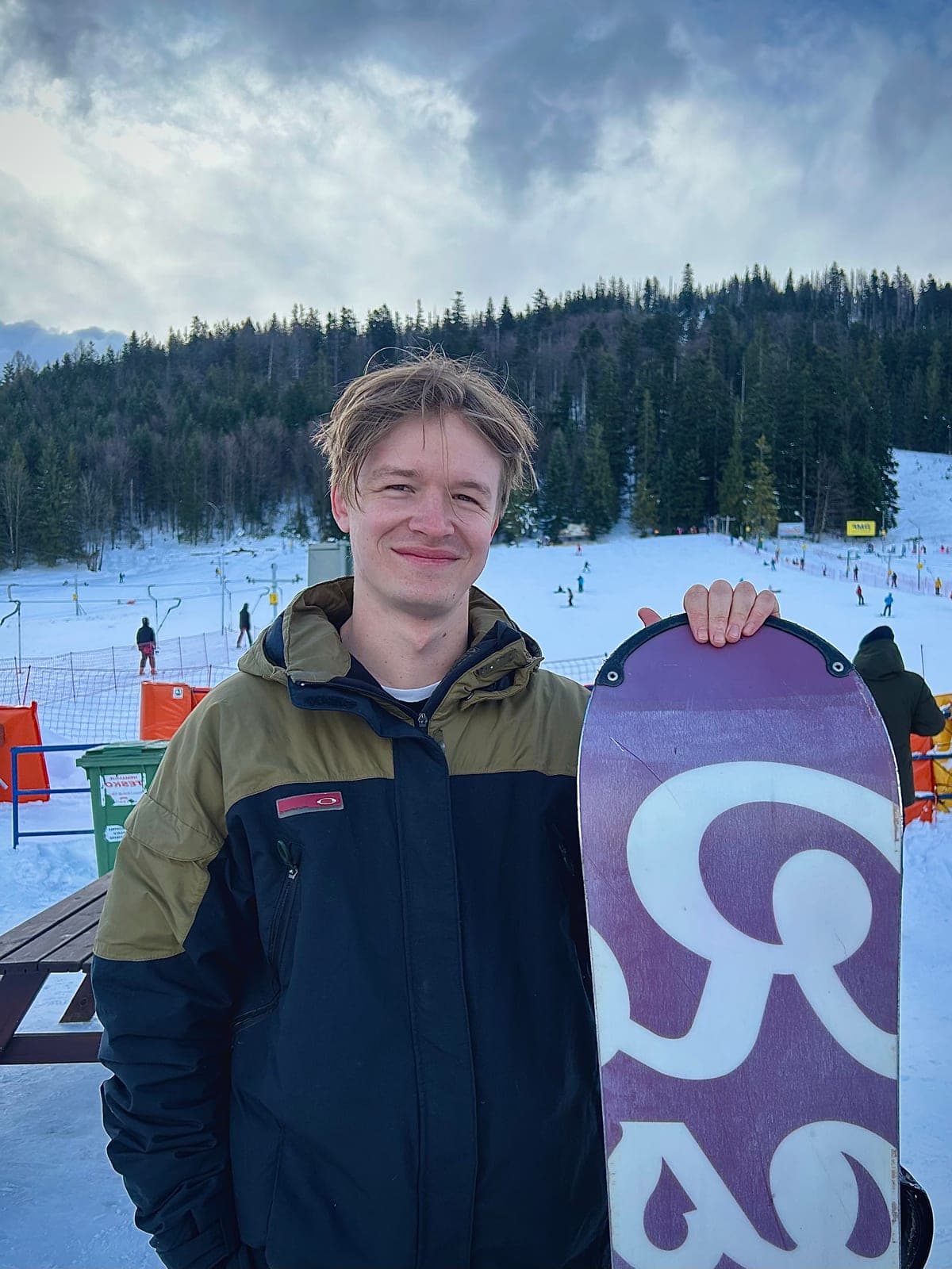 Matt Botha, smiling, holding a snowboard on a ski slope with pine forest behind him.